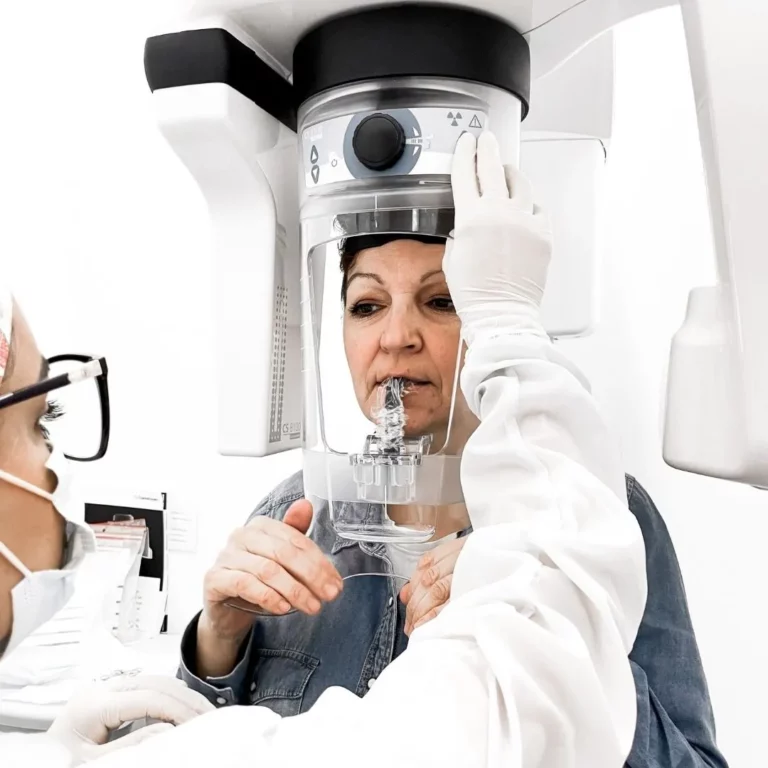 Mulher posicionada num aparelho de radiografia dentária, acompanhada por uma profissional.