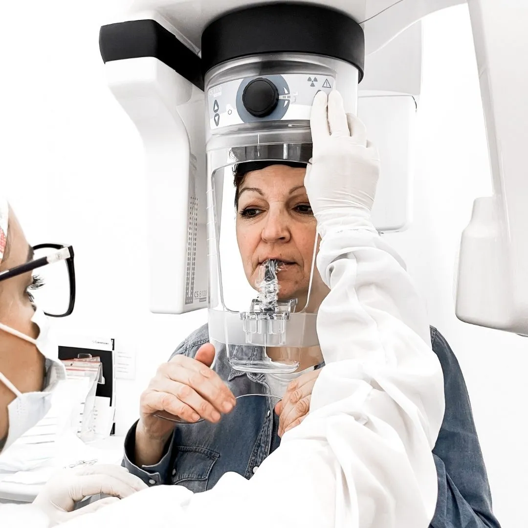 Mulher posicionada num aparelho de radiografia dentária, acompanhada por uma profissional.