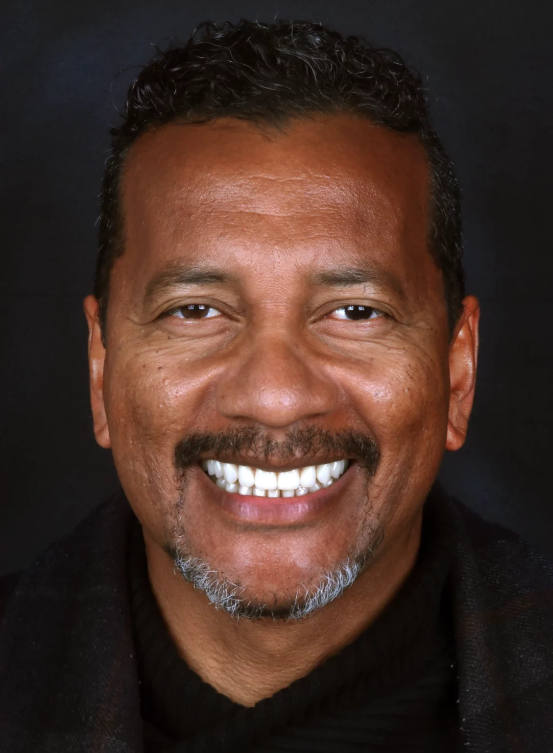 Retrato frontal de um homem de bigode a sorrir, com os dentes visíveis.