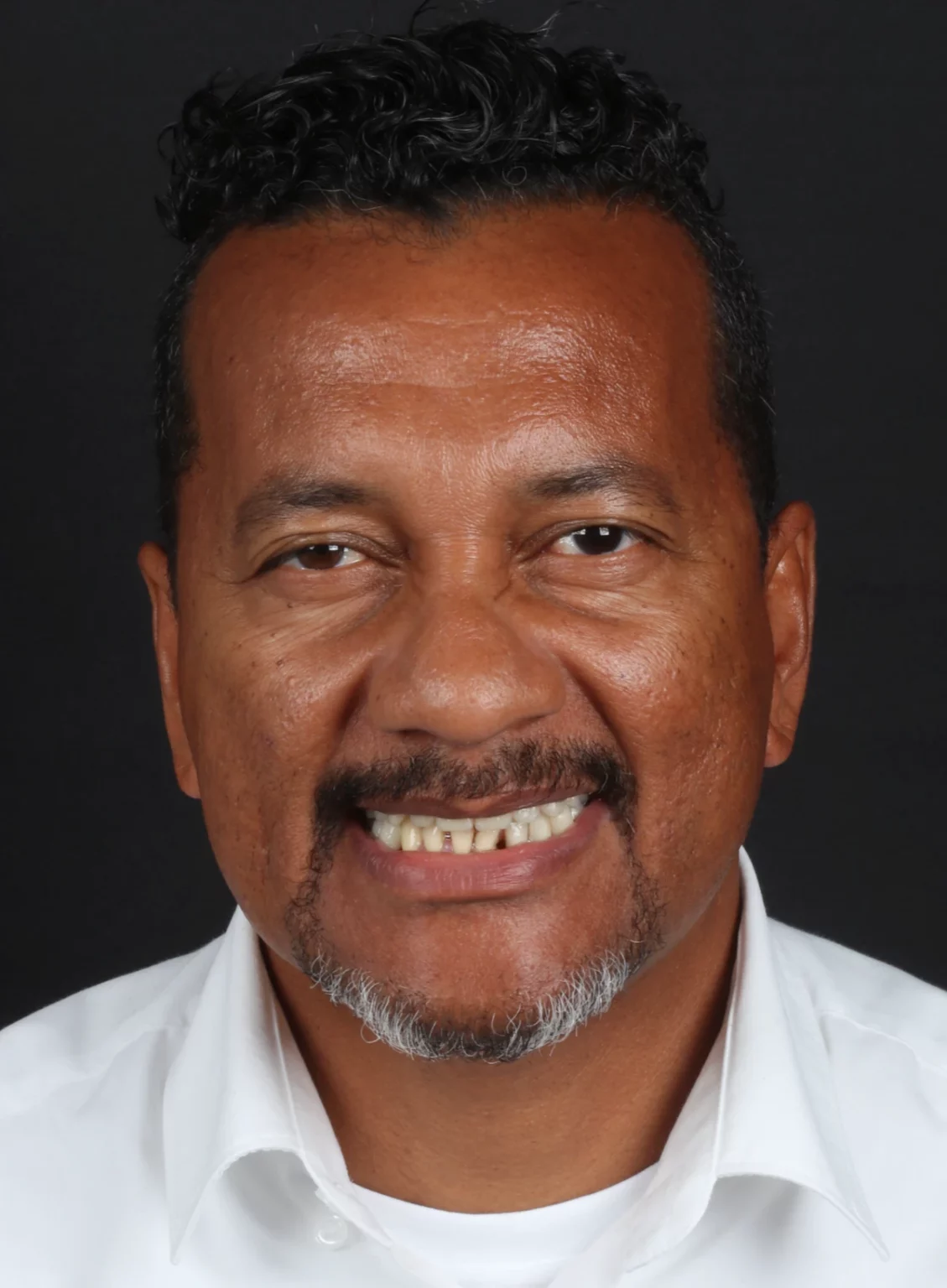 Retrato frontal de um homem de bigode e camisa branca a sorrir.
