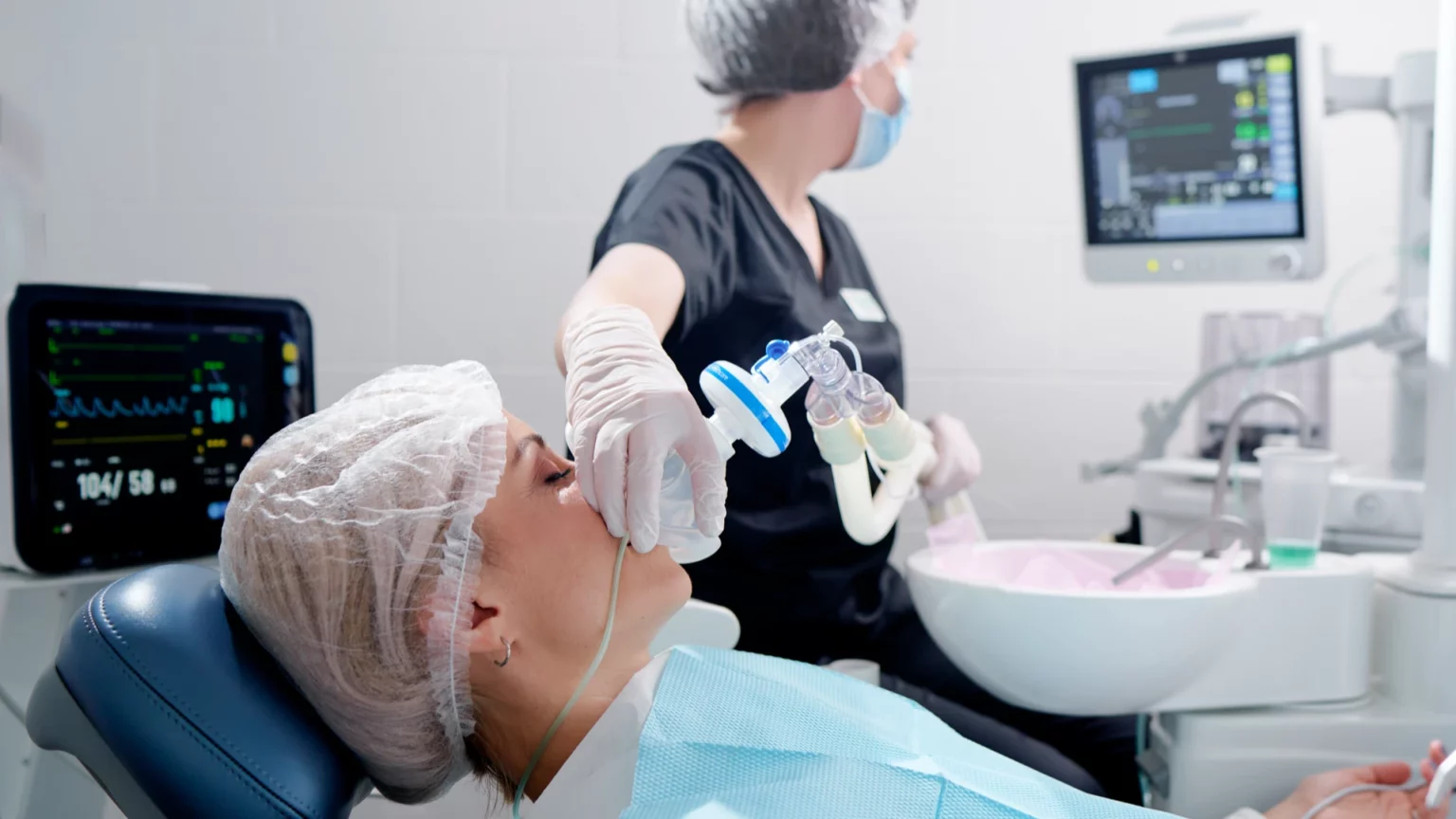 Pessoa com máscara na cadeira dentária, acompanhada por uma profissional junto a monitores.